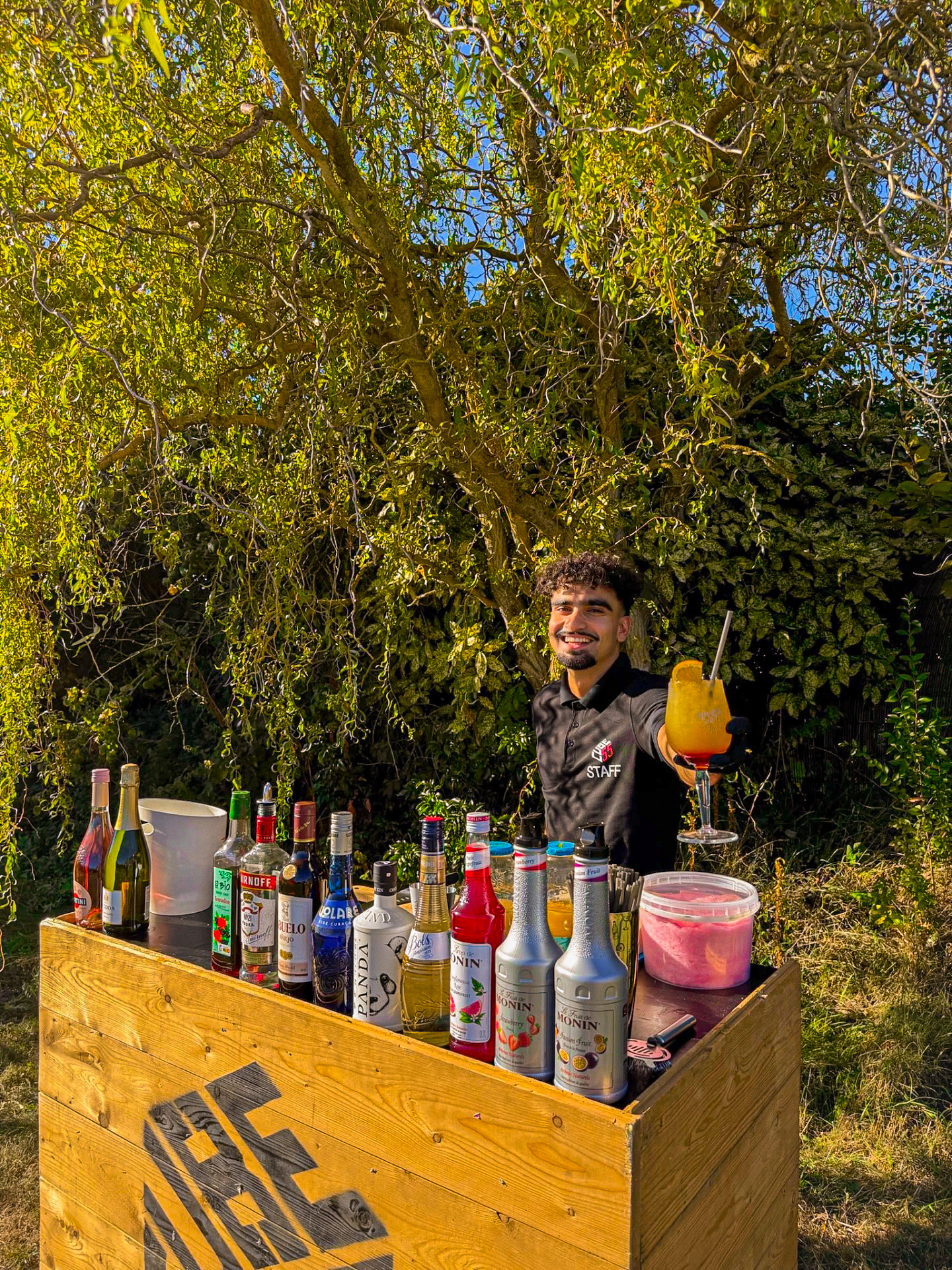 Barman présentant un cocktail orange derrière son bar mobile en bois dans un jardin
